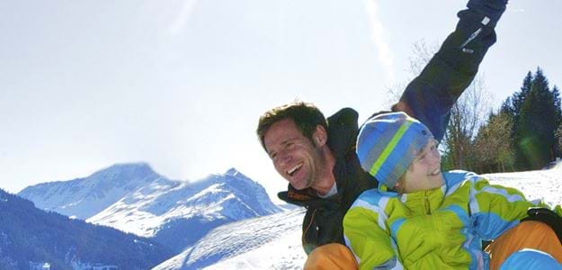 Far og søn leger i sneen med udsigt til bjerge i Saalbach